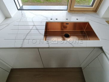 Realizace pracovní desky z technistone Crystal Calacatta Silva zahrnující měděný dřez se spodní montáží, vyfrézované odkapové drážky a výřezy pro baterii a dávkovač saponátu. Kamenný parapet je součástí kuchyňské desky. Zakázková výroba TONE OF STONE.