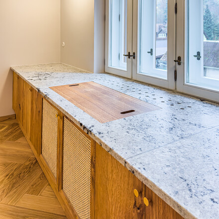 Kuchyňsk&aacute; pracovn&iacute; deska z indick&eacute; žuly Colonial White v &uacute;pravě sat&eacute;n. Do kuchyňsk&eacute; desky je vsazen dřez se spodn&iacute; mont&aacute;ž&iacute; a zabudovan&yacute; projektor. Integrovan&yacute; parapet s průduchy pro prouděn&iacute; tepl&eacute;ho vzduchu z topen&iacute; je souč&aacute;st&iacute; pracovn&iacute; desky. Zak&aacute;zkov&aacute; v&yacute;roba TONE OF STONE.     